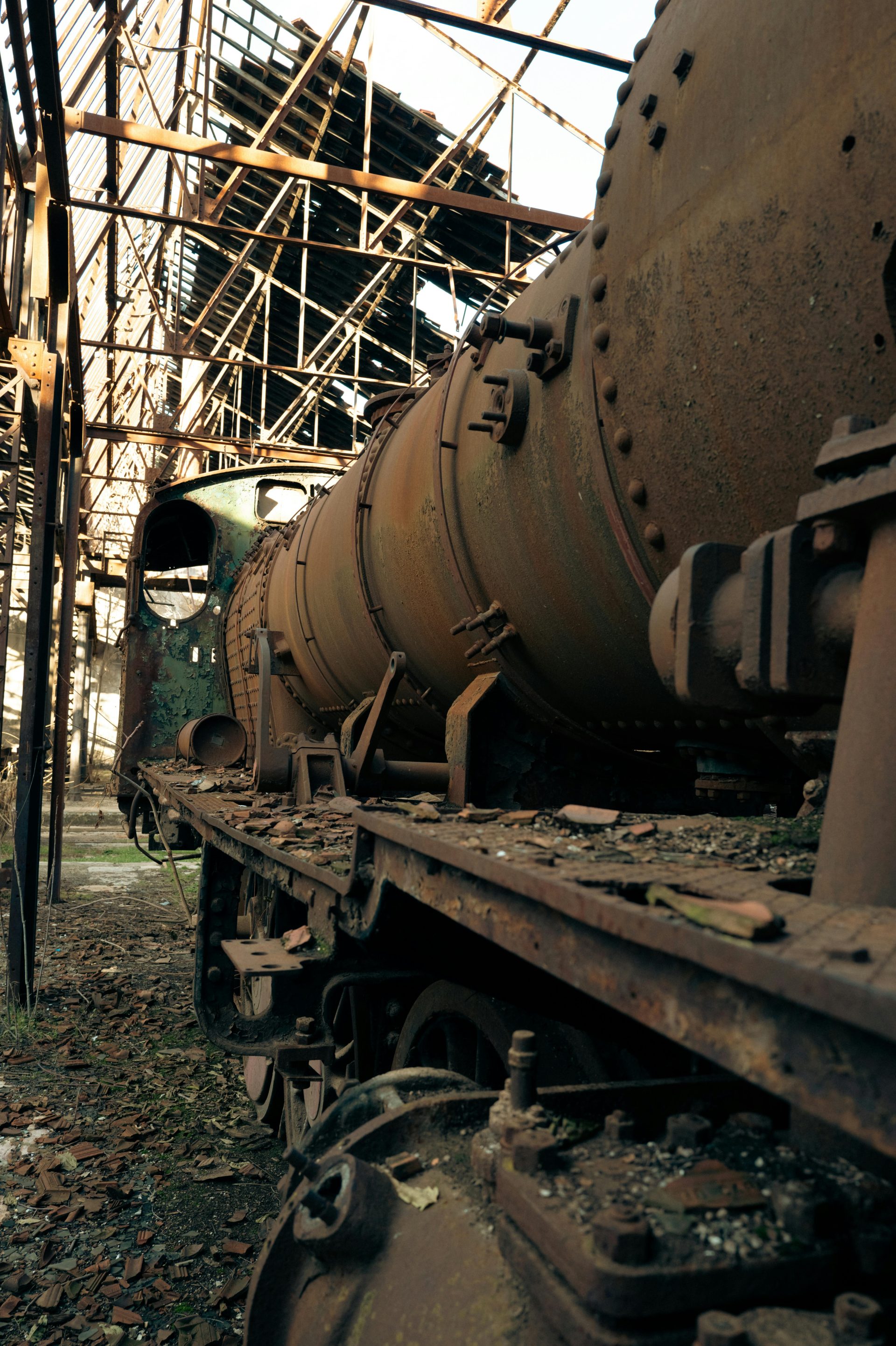 A rusted obsolete train inside a deteriorating industrial site, with sunlight filtering through broken roofs.